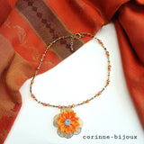 collier artisanal composé de perles dorées et orangées, orné d'un pendentif fleur en satin orange sur support métallique doré, présenté sur un tissu orange