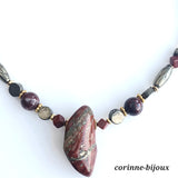 Un colllier en pierres fines, aux couleurs naturelles composé de jaspe rouge, une pierre de couleur brique rouge, mélé à une pierre fine appelée pyrite, une pierre de couleur brillant opaque. L’ensemble de ces pierres se retrouve dans la couleur de la pierre centrale du collier appelé jaspe