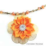 Gros plan du pendentif du collier représentant une fleur en tissu satin orange avec un centre orné de strass, l'ensemble monté sur des pétales en résine dorées.