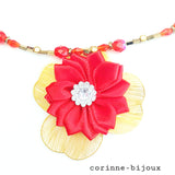 Gros plan du pendentif du collier représentant une fleur en tissu satin rouge avec un centre orné de strass, l'ensemble monté sur des pétales en résine dorées.