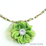 Gros plan du pendentif du collier représentant une fleur en tissu satin vert avec un centre orné de strass, l'ensemble monté sur des pétales en résine dorées.